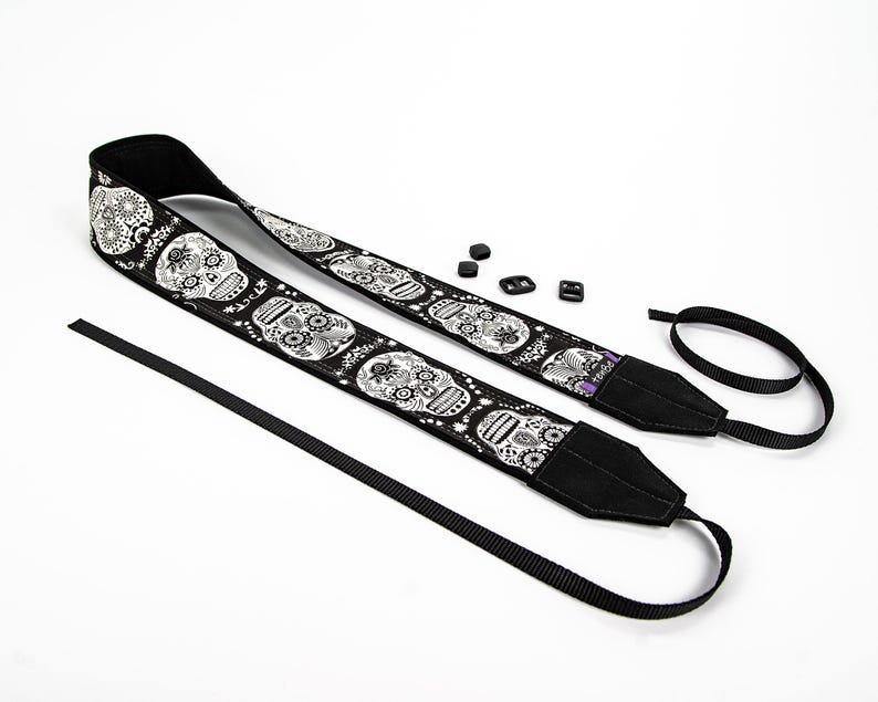 Black and white camera strap with a repeating pattern of sugar skulls. The skulls are intricately detailed with floral designs. The strap is made of a durable fabric and features a comfortable, adjustable design.  Perfect for photographers who appreciate unique and stylish accessories.