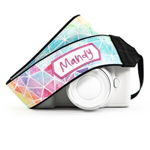 May include: A camera strap featuring a colorful geometric watercolor pattern with white lines and a rectangular patch embroidered with the name Mandy in pink thread, attached to a white camera body with black fabric ends and black trim.