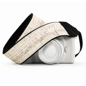 May include: A white camera with a musical note patterned strap. The strap is cream-colored with brown musical notation and black backing. The camera is white with a large lens and a black strap attachment.
