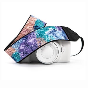 May include: A white digital camera with a colorful patterned strap. The strap features a design with shades of purple, teal, and peach. The camera strap is made of a durable material and is designed to be comfortable.