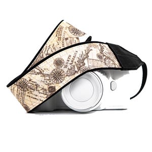 May include: A camera strap with a vintage map design. The strap features a cream-colored background with a detailed map print, compass roses, and black accents. The strap is wrapped around a white camera body.