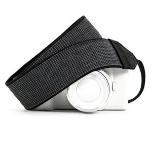 May include: A black and gray houndstooth camera strap draped over a white camera body. The strap is made of a woven fabric with a repeating pattern. The camera has a silver lens and a black strap attachment.