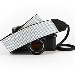 May include: A black Canon camera with a 50mm lens attached, featuring a wide camera strap with a white and blue houndstooth pattern. The strap has black fabric ends and is draped over the top of the camera body, which displays the text LENS MADE IN JAPAN.