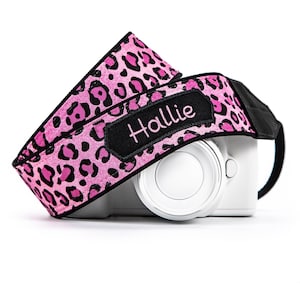 May include: A camera strap featuring a vibrant pink and black leopard print pattern. The strap includes a black rectangular patch with the name Hallie embroidered in pink thread, resting on a white camera body with a circular lens mount.