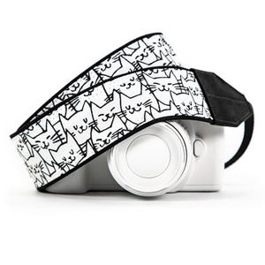 May include: A white digital camera with a black and white camera strap featuring a repeating pattern of hand-drawn cat faces. The strap has black edges and solid black end pieces, while the camera body is plain white with a circular lens.