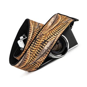 May include: A brown and black camera strap with a pheasant feather pattern. The strap is made of a soft, padded material and is designed to be comfortable to wear.