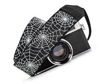 May include: A black camera strap featuring a repeating white spiderweb pattern attached to a black and silver camera. The strap is made of fabric and displays intricate web designs across its entire length, contrasting with the camera body.