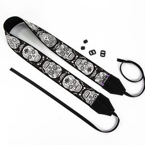 Black and white camera strap with a repeating pattern of sugar skulls. The skulls are intricately detailed with floral designs. The strap is made of a durable fabric and features a comfortable, adjustable design.  Perfect for photographers who appreciate unique and stylish accessories.