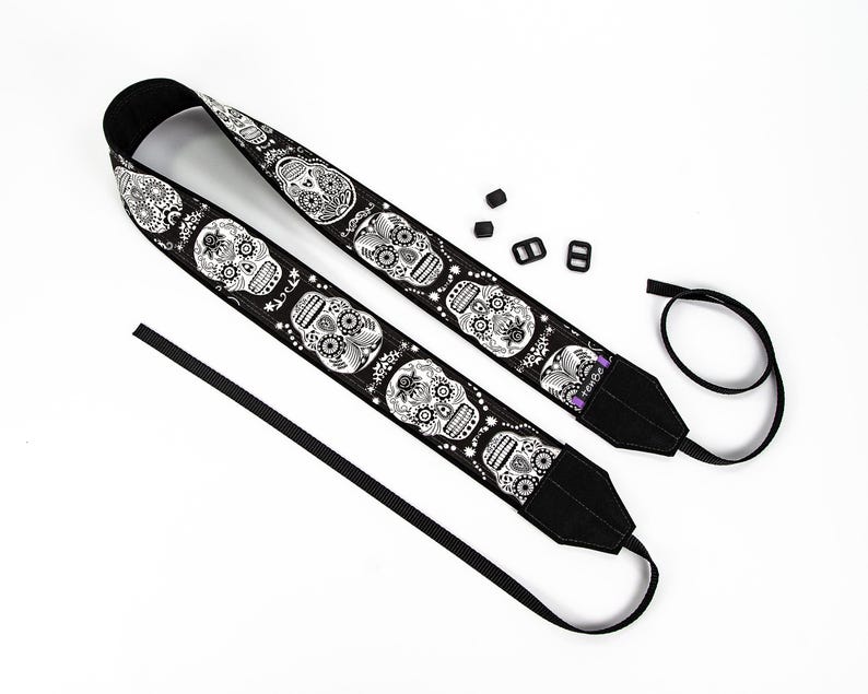 Black and white camera strap with a repeating pattern of sugar skulls. The skulls are intricately detailed with floral designs. The strap is made of a durable fabric and features a comfortable, adjustable design.  Perfect for photographers who appreciate unique and stylish accessories.