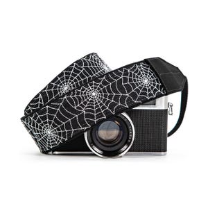 May include: A black camera strap featuring a repeating white spiderweb pattern draped over a black and silver digital camera. The strap has a wide, flat design with solid black fabric ends and a visible lens on the front of the camera body.