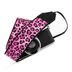 Puede incluir: Correa de cámara de leopardo rosa y negro con una cámara negra.