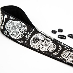 Black and white camera strap with a repeating pattern of sugar skulls. The skulls are intricately detailed with floral designs. The strap is made of a durable fabric and features a comfortable, adjustable design.  Perfect for photographers who appreciate unique and stylish accessories.