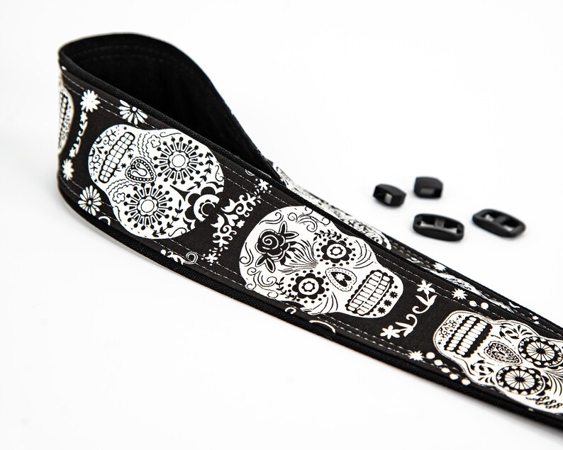 Black and white camera strap with a repeating pattern of sugar skulls. The skulls are intricately detailed with floral designs. The strap is made of a durable fabric and features a comfortable, adjustable design.  Perfect for photographers who appreciate unique and stylish accessories.