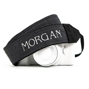 May include: A gray and black houndstooth camera strap with the name "MORGAN" embroidered in white. The strap is draped over a white camera body, with a black strap connector.