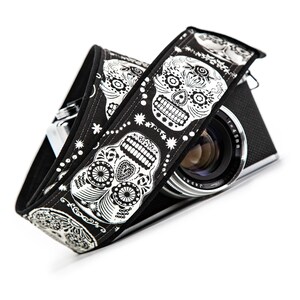 Black and white camera strap with a repeating pattern of sugar skulls. The skulls are intricately detailed with floral designs. The strap is made of a durable fabric and features a comfortable, adjustable design.  Perfect for photographers who appreciate unique and stylish accessories.
