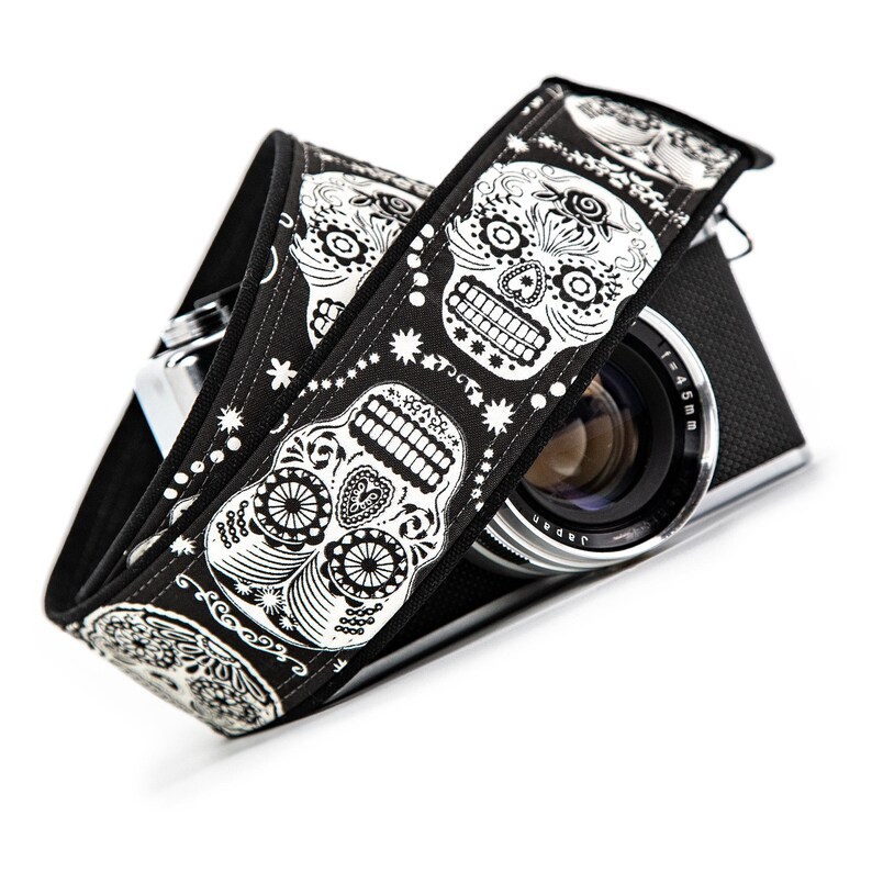Black and white camera strap with a repeating pattern of sugar skulls. The skulls are intricately detailed with floral designs. The strap is made of a durable fabric and features a comfortable, adjustable design.  Perfect for photographers who appreciate unique and stylish accessories.