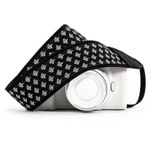 May include: A black camera strap with a white fleur-de-lis pattern is draped over a white digital camera. The strap is made of a durable fabric and features a comfortable design for carrying a camera. The camera has a silver lens.