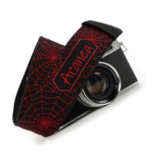 May include: Black camera strap with red spiderweb design and the name "Aranea" embroidered in red. The strap is wrapped around a black and silver camera with a lens.