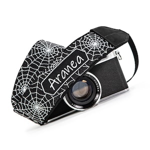 May include: Black and white camera strap with a spiderweb design. The strap has the word "Aranea" embroidered on a black label. A black and silver camera is partially visible behind the strap.