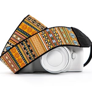 May include: A white digital camera with a patterned camera strap featuring a repeating design of orange spirals, geometric triangles, and diamonds in shades of yellow, green, blue, and black, finished with black fabric ends and black trim.