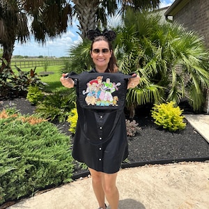 May include: Black button-down dress with a colorful embroidered image of Toy Story characters. The dress has a shoulder strap and is held up by a person. The person is wearing sunglasses and mouse ears.