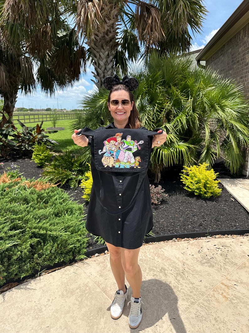 May include: Black button-down dress with a colorful embroidered image of Toy Story characters. The dress has a shoulder strap and is held up by a person. The person is wearing sunglasses and mouse ears.