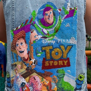 May include: A light blue acid-wash denim vest featuring a large, colorful Toy Story graphic on the back. The design includes Buzz Lightyear, Woody, and other characters, embellished with numerous small, sparkling rhinestones throughout the image.