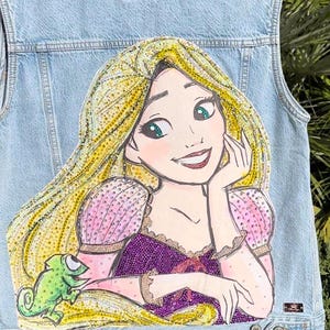 Puede incluir: Un chaleco vaquero azul claro con una imagen de lentejuelas de Rapunzel de la película de Disney Enredados. Rapunzel lleva un vestido morado y rosa y tiene el pelo largo y rubio. Un camaleón verde está en su pelo.