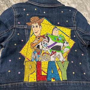 May include: A blue denim jacket with a yellow star design featuring the characters Woody, Buzz Lightyear, and Jessie from the Toy Story franchise. The star has rhinestones and the text "Made to Play" in colorful rhinestones.