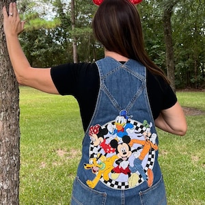 May include: Denim overall shorts with a colorful Mickey Mouse and friends graphic on the back. The graphic features Mickey, Minnie, Donald, Goofy, and Pluto. A pair of red sequined mouse ears are worn on the head.