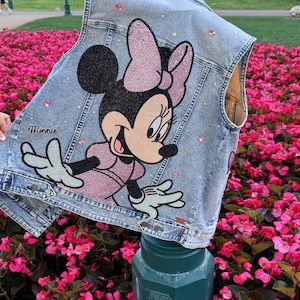 May include: A light blue denim vest featuring a large, rhinestone-embellished graphic of Minnie Mouse wearing a pink dress and bow. The design includes the name Minnie in script, scattered pink and clear rhinestones, metal buttons, and seams.