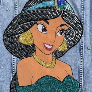 May include: A light blue denim jacket featuring a large, detailed appliqué of a character with black hair, a teal headband with a blue jewel, gold earrings, and a gold necklace. The character wears a teal off-the-shoulder top, all embellished with rhinestones.