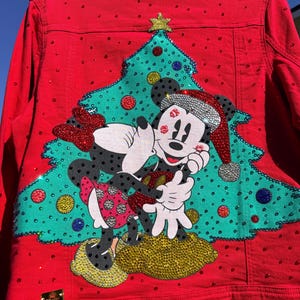 May include: Red denim jacket with a Christmas tree and Mickey and Minnie Mouse design. The jacket is embellished with rhinestones and features a gold emblem. The Christmas tree is green with colorful ornaments.