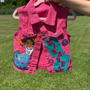 May include: A pink denim vest with silver buttons and rhinestone accents. The vest features an embroidered image of a character and the name "MIRABEL" on patches. The vest is hanging on a pink hanger.