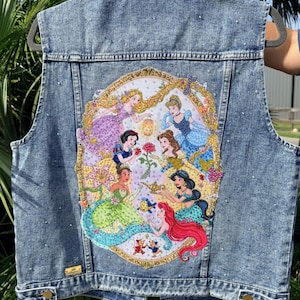 May include: A blue acid-wash denim vest featuring a large, oval-shaped embroidered patch on the back. The patch depicts seven animated princesses in colorful gowns arranged around a central rose, a golden lamp, and small mice, all framed in gold.
