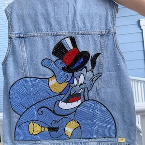 May include: A light blue denim vest featuring a large, detailed graphic of a blue genie character on the back. The design is embellished with numerous small, sparkling rhinestones in various colors, including red, black, gold, and blue, forming the genie's body and top hat.