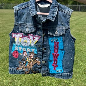 May include: A denim vest with a button-up front, embellished with rhinestones. The vest features a "Toy Story" patch with Woody and Buzz Lightyear, and a "Hey Howdy Hey" patch in blue and pink. The vest is held up by a pink hanger.