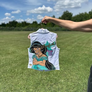 May include: White denim vest featuring a colorful illustration of Princess Jasmine from Aladdin. The artwork includes the text "Magical!" and a depiction of a palace. The vest is held up against a backdrop of a green field and blue sky.