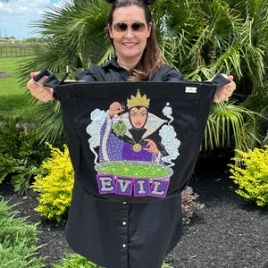 May include: Black tote bag featuring a colorful graphic of a cartoon queen with a crown, holding a green apple over a bubbling cauldron. The word "EVIL" is spelled out in purple letters below the cauldron. The bag has black handles.