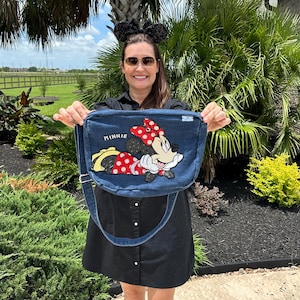 May include: A denim messenger bag featuring an embroidered Minnie Mouse design with a red polka dot bow. The bag has a long adjustable strap and the word "MINNIE" is embroidered on the bag. The bag is held up in front of a green background.