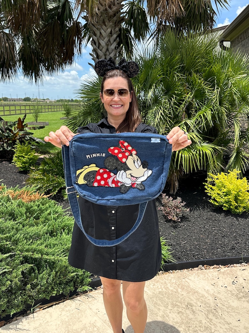 May include: A denim messenger bag featuring an embroidered Minnie Mouse design with a red polka dot bow. The bag has a long adjustable strap and the word "MINNIE" is embroidered on the bag. The bag is held up in front of a green background.