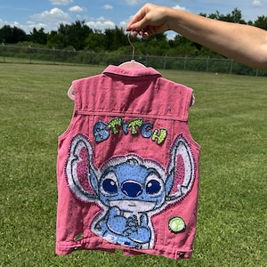 May include: Pink denim vest featuring a large, embroidered image of the character Stitch from the Disney movie Lilo & Stitch. The word "Stitch" is embroidered in green and yellow. The vest is decorated with small, sparkling embellishments.