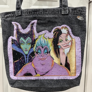 May include: A charcoal gray denim tote bag featuring a rectangular patch with illustrations of three animated villains. The patch is bordered by light purple fabric with visible stitching and small white beads, accented with colorful rhinestones.