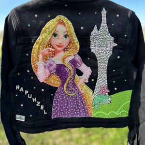 May include: Black denim jacket with a rhinestone design featuring Rapunzel from Tangled. The design includes Rapunzel, her tower, and Pascal the chameleon.