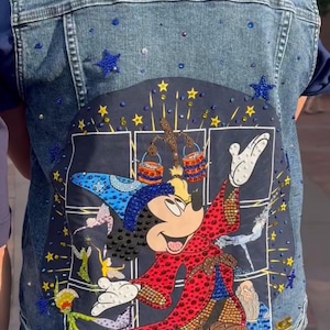 May include: A blue denim vest with a large embroidered patch featuring Mickey Mouse as a sorcerer. The patch is covered in rhinestones and depicts Mickey Mouse with a wand and a hat, surrounded by stars and other magical elements.