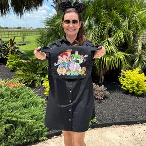 May include: Black crossbody bag featuring a colorful graphic of Disney's Toy Story characters. The bag has a long strap and is held up by a person. The background includes green plants and a sunny sky.