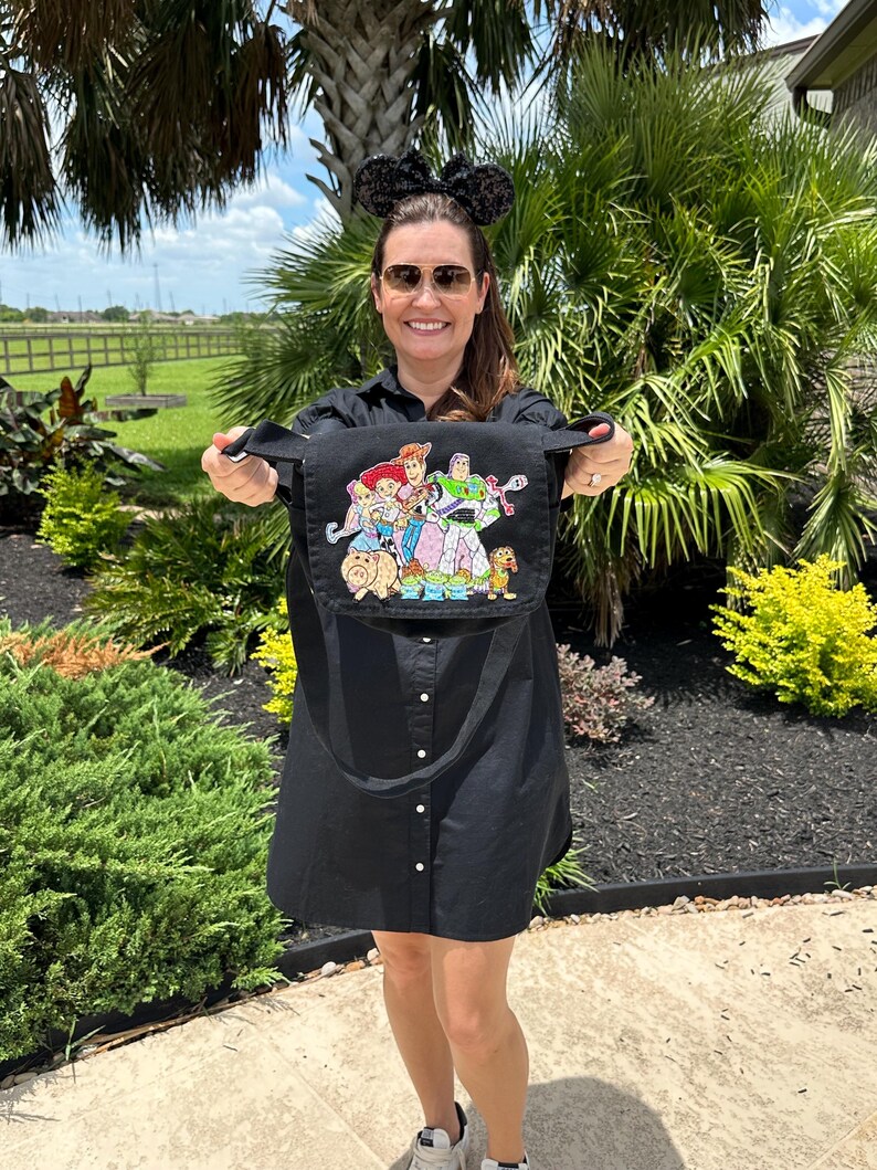May include: Black crossbody bag featuring a colorful graphic of Disney's Toy Story characters. The bag has a long strap and is held up by a person. The background includes green plants and a sunny sky.