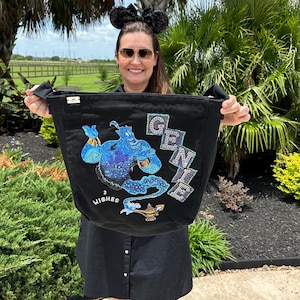 May include: Black tote bag featuring a sparkling blue Genie design with the text "GENIE" and "3 WISHES". The bag has black handles and is held up in front of a person. The background includes palm trees and green foliage.