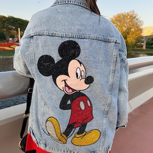 May include: A light blue denim jacket featuring a large, beaded Mickey Mouse character on the back. The design includes a black head, red shorts, and yellow shoes, all rendered in small beads. The jacket fabric is embellished with scattered rhinestones.
