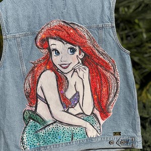 May include: A light blue denim vest with a rhinestone embellished image of Ariel, the Little Mermaid, on the back.